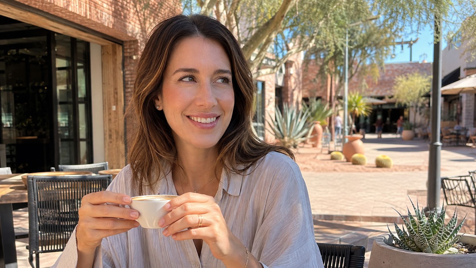 A woman smiling confidently while holding a coffee cup at an outdoor cafe with desert landscape scenery.